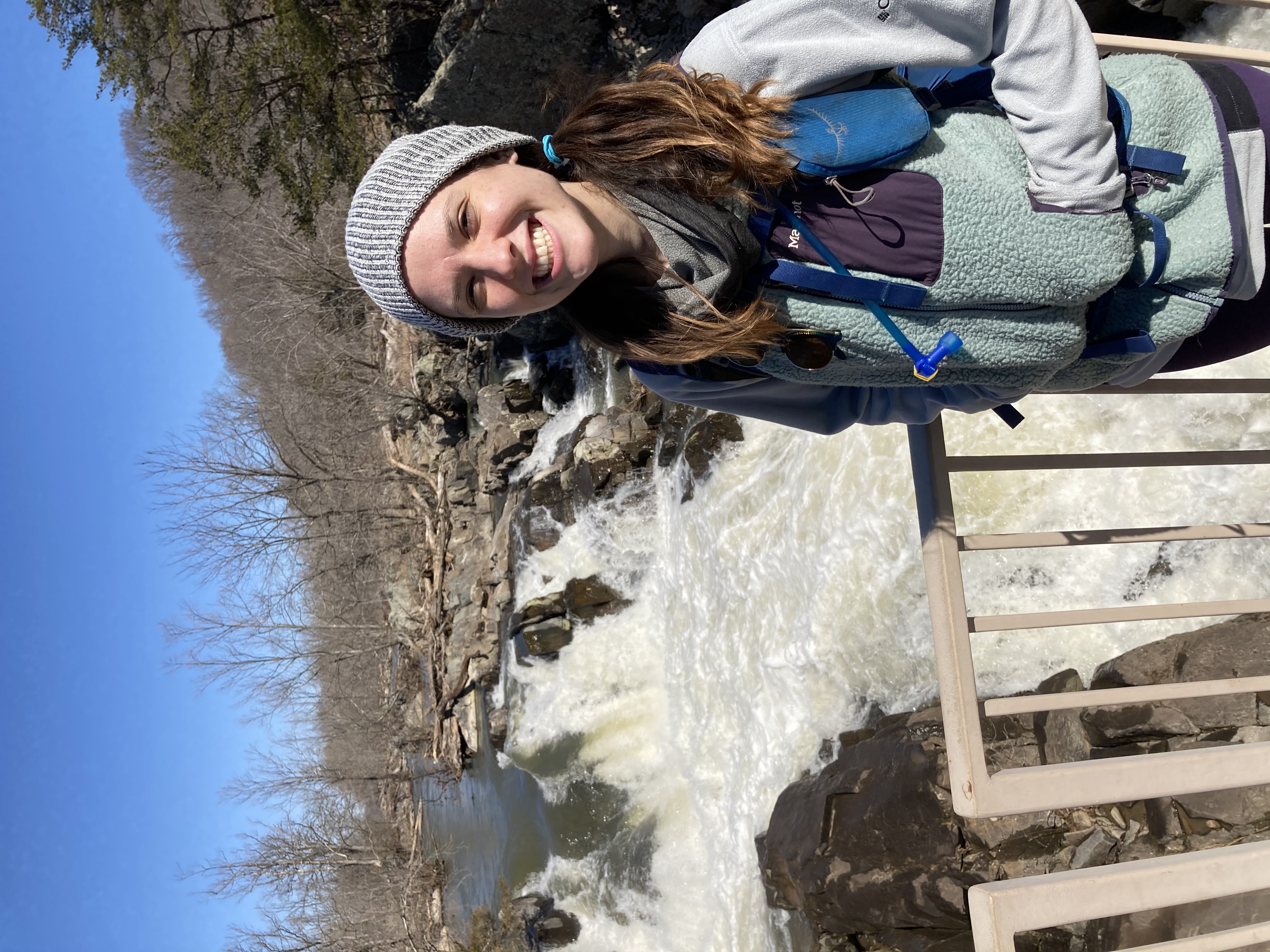 Rebecca Deegan smiling and wearing a knit hat while hiking along a swift moving creek on a chilly day.