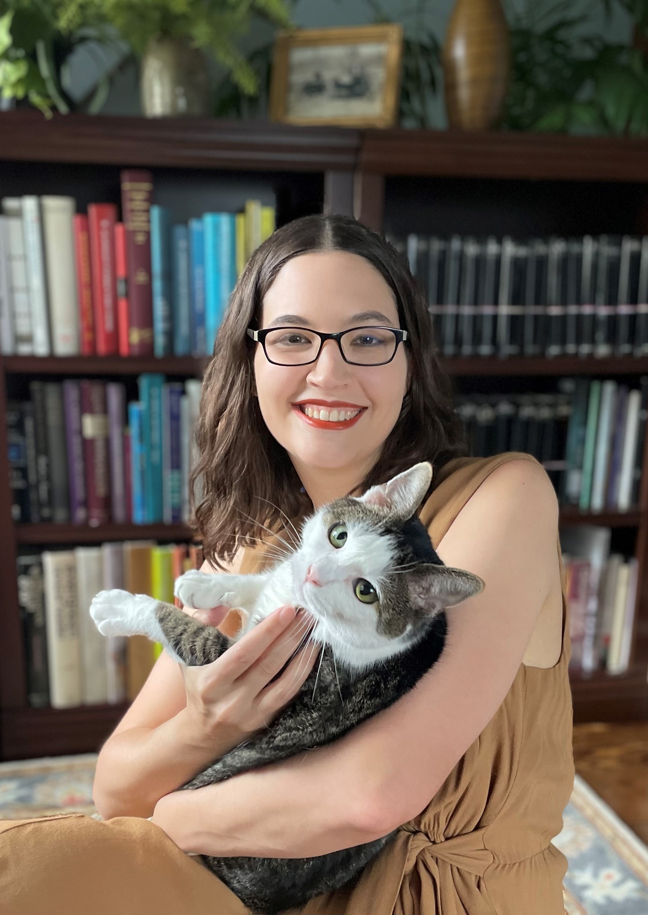 Melissa Farr is smiling and holding her brown and white cat in front of a bookshelf with plants. She has brown wavy hair, glasses, and is wearing a tan jumpsuit.