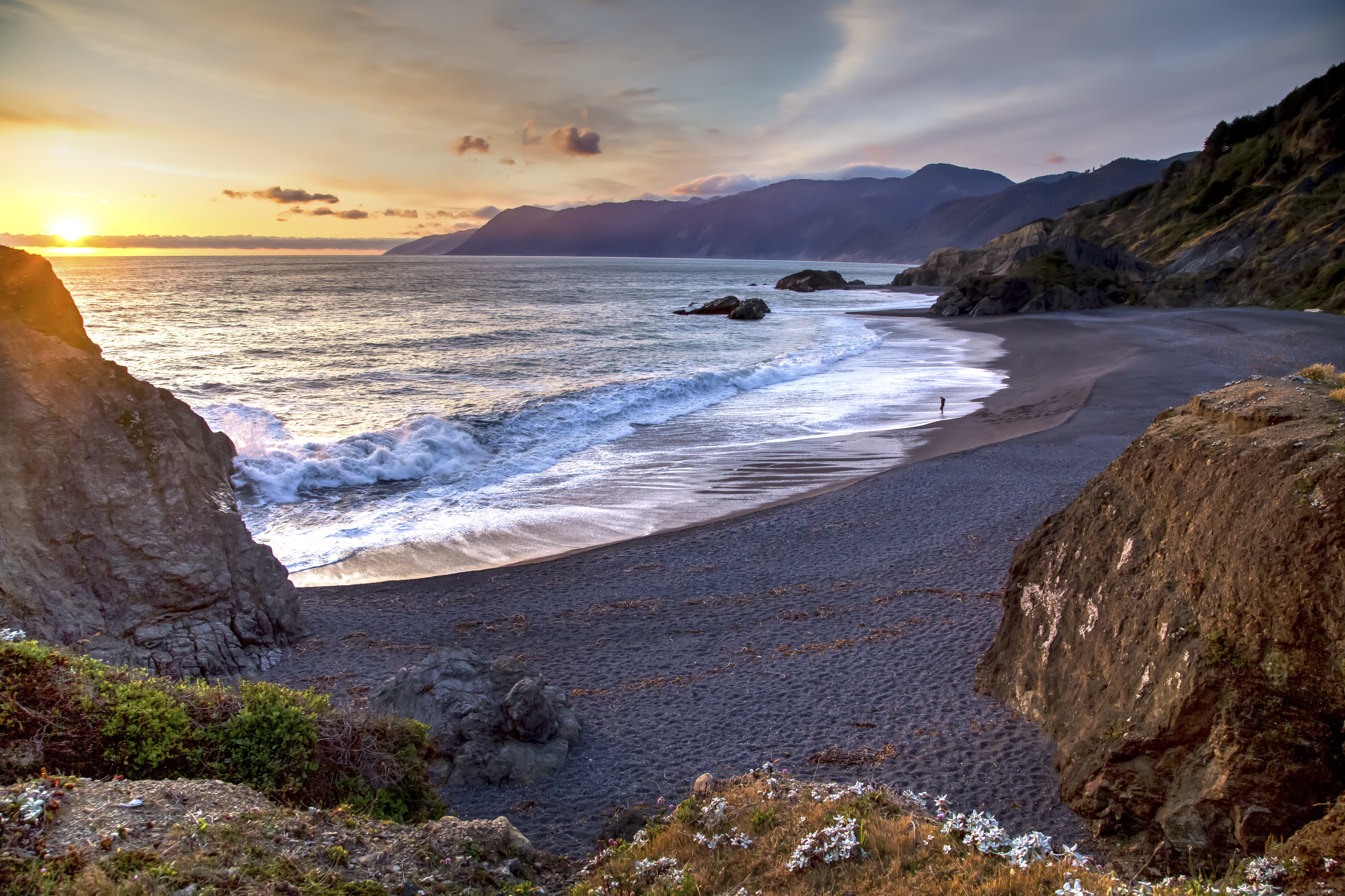 Shallow ocean waves backed by an orange setting sun in the distance wash up onto a curved sandy beach surrounded by large rocks which extend into mountains in the distance.