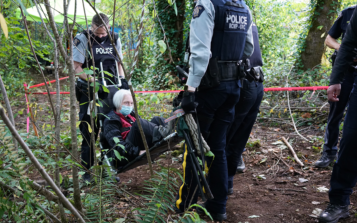 Gray-haired protestor in red is carried through a forest by police officers removing her from a protest.
