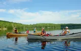 Four adults and two children travel in two canoes across a calm lake with trees in the background