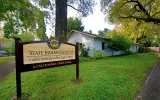  A sign reading "State Indian Museum California Indian Heritage Center / A California State Park" sits in front of a one-story painted-white adobe building with trees surrounding it.