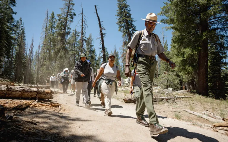 Shelton Johnson, wearing a park ranger uniform, walks down a dusty path through woods followed by a dozen or so hikers.