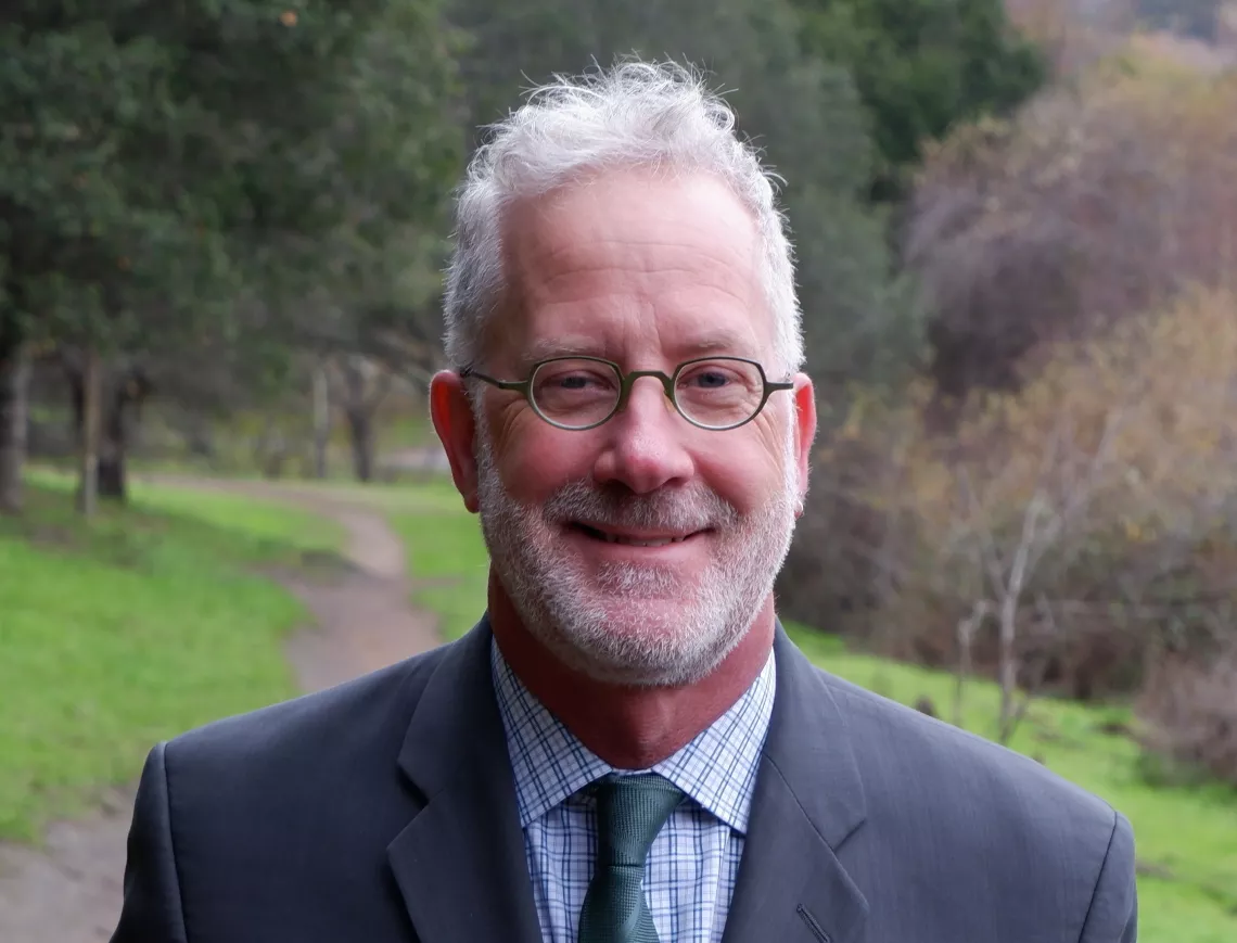 Nick Pilch wears a gray suit and smiles on a green natural backdrop