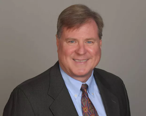 Joe Kovalick smiles against a gray background wearing a black suit.