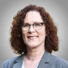 A photo of woman with curly, shoulder length chestnut-colored hair and glasses smiles at the camera. She is pictured from the shoulders up and is wearing a gray blazer and white shirt. She is framed by a gray background.
