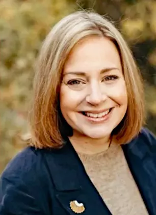 A photographic portrait woman with shoulder length blond hair smiling at the camera. She is wearing a blue blazer and tan shirt, and is framed by a blurry backdrop of fall leaves. 