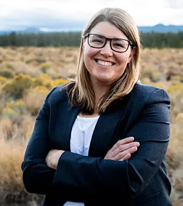 A portrait photo of a woman from the waist up. She has blonde, shoulder-length hair, glasses, and is standing with her arms crossed in front of her. She is wearing a black blazer and a white shirt. She is in the middle of a natural field with yellow bushes, and in the distance are a dark green forest and blue mountains.