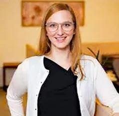 A woman with reddish-blonde hair and glasses smiles at the camera. She is pictured from the waist up and is wearing a black shirt and white cardigan. She stands against an abstract background of yellow furniture and a painting.
