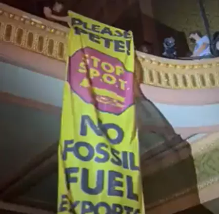 A long yellow banner reading "Please Pete! STOP S.P.O.T. No Fossil Fuel Exports!" is unfurled from a balcony.
