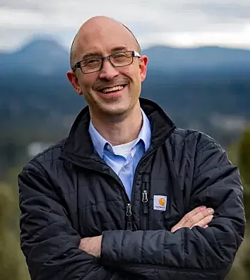 A mostly bald man with glasses stands, pictured from the waist up, smiling broadly at the camera with his arms crossed. He is wearing a black jacket over a light blue button down shirt. He stands against a blurry backdrop of blue mountains in the distance and light green farmland in the foreground.