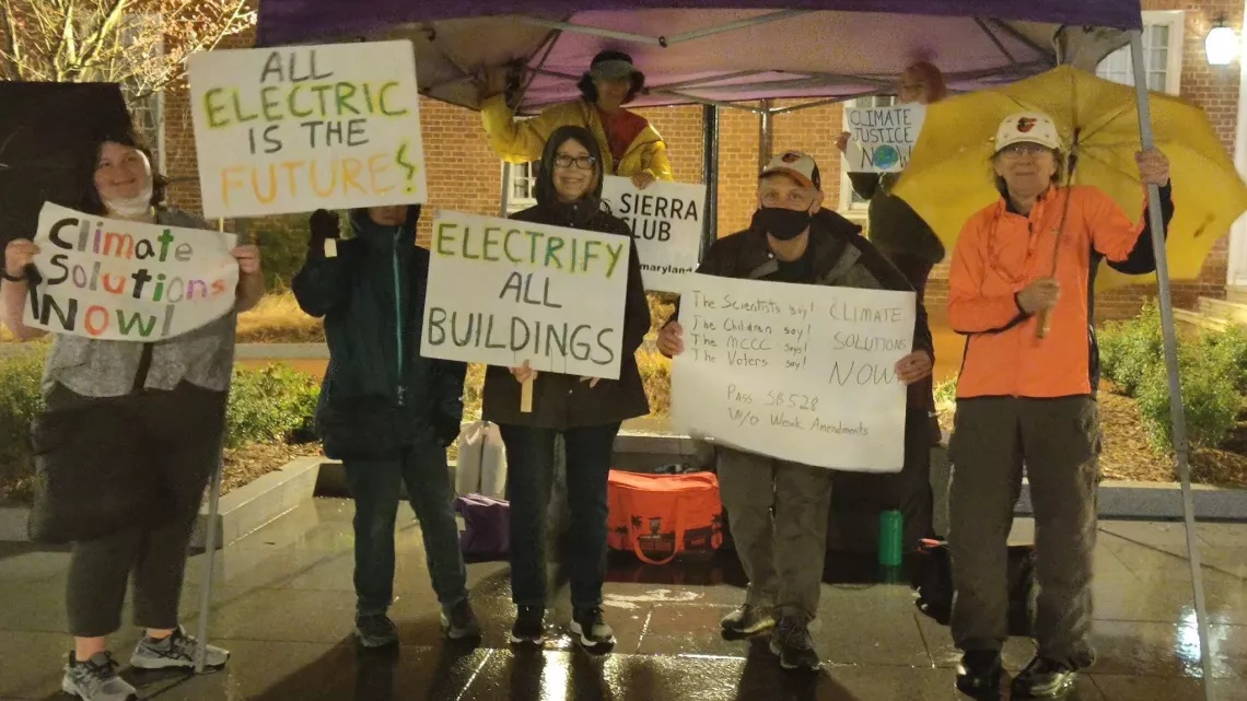 Volunteers gather under tent on rainy day holding building electrification signs