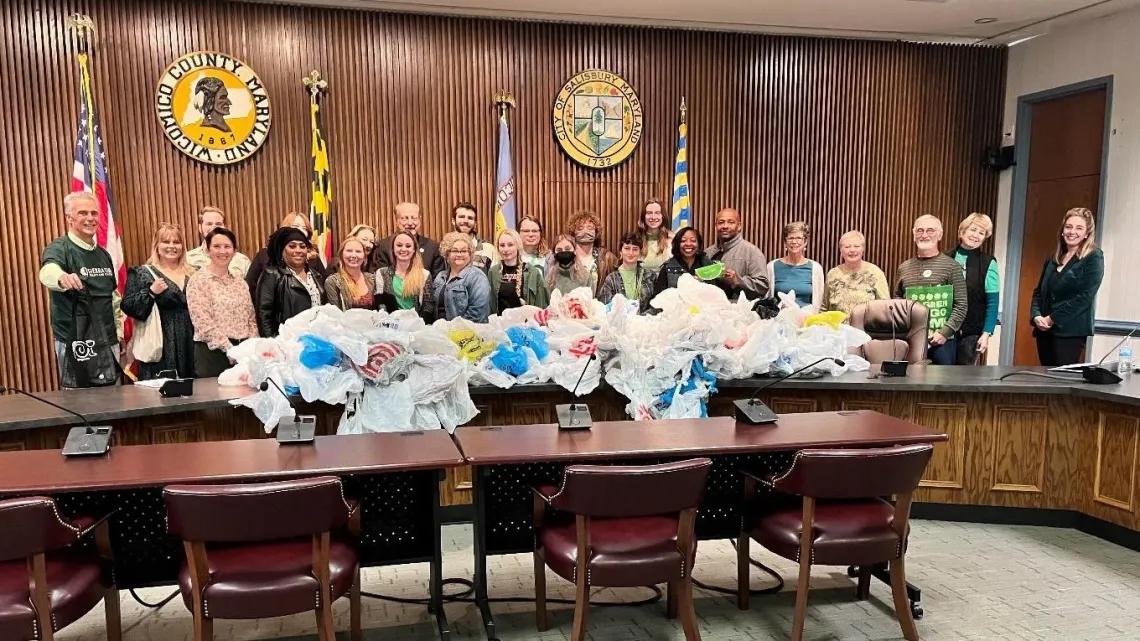 A dozen people gather in council chambers in front of a table with plastic bags.