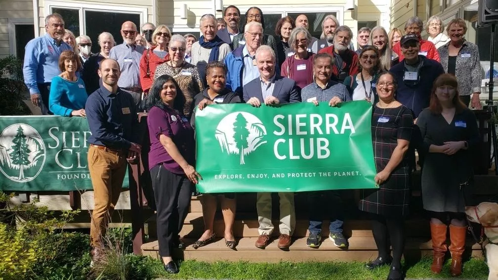 A group of volunteers and elected officials gather in front of Sierra Club sign outside 