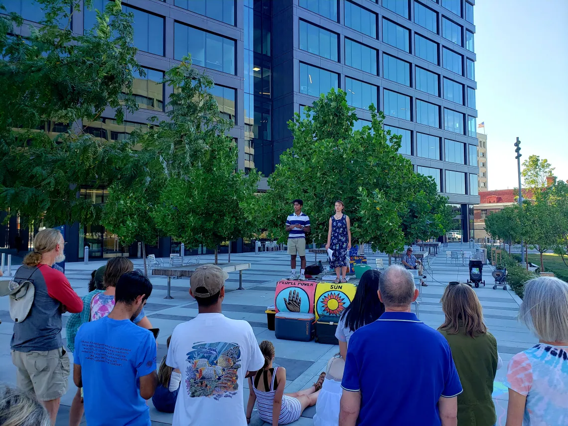 Picture of two high school students standing on an outdoor table with banners on it speaking to a crowd of people listening