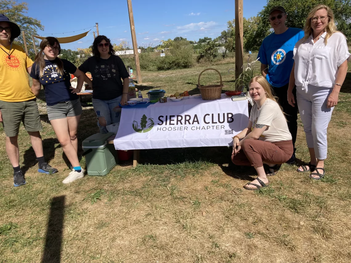 A group of six people outdoors on a blue-sky day, standing around a picnic table with a white banner with the Sierra Club Hoosier Chapter logo on it. They are all smiling and looking at the camera.