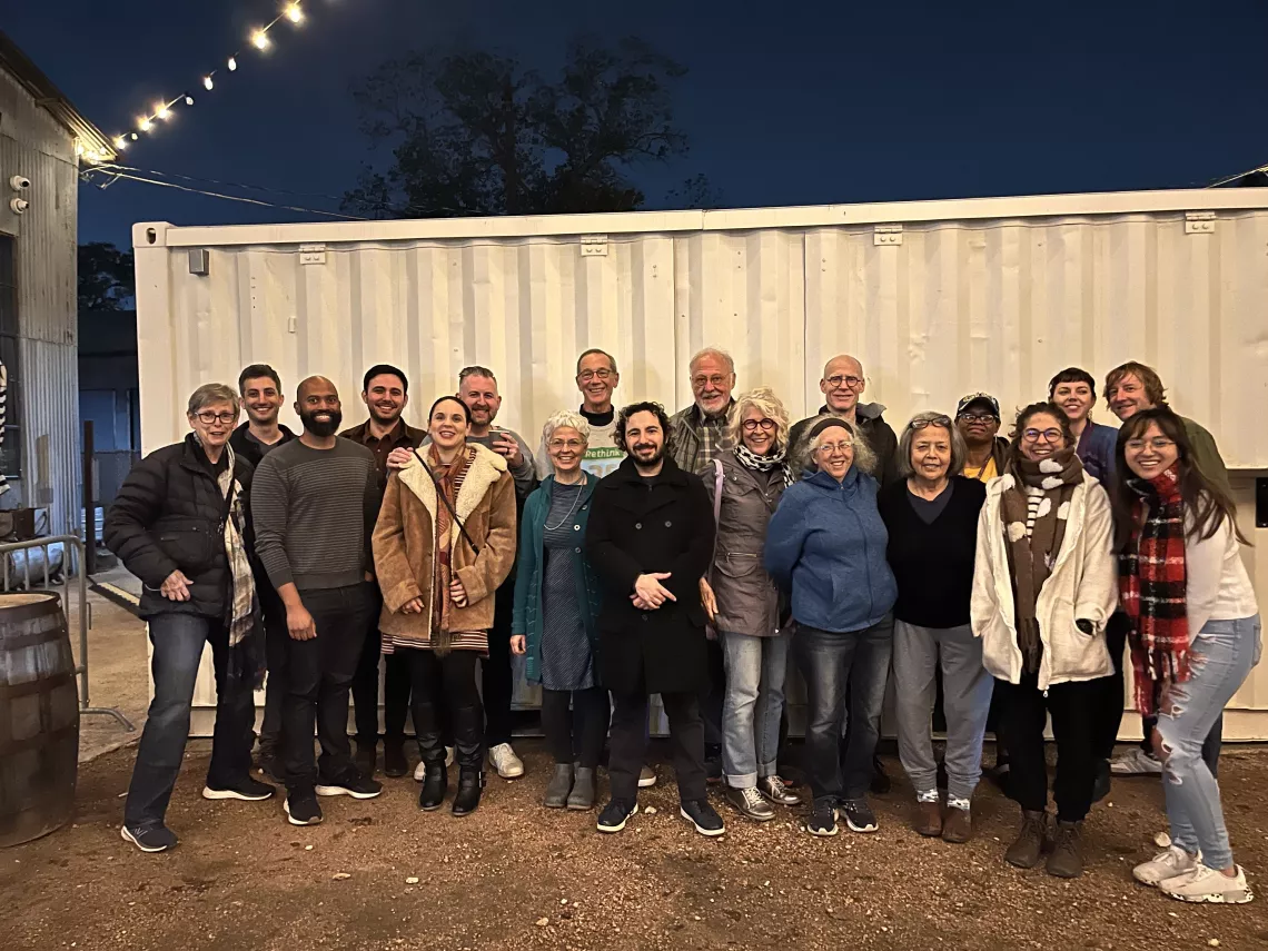 A multigenerational, multiracial group of folks poses for a picture in an outdoor courtyard at dusk.