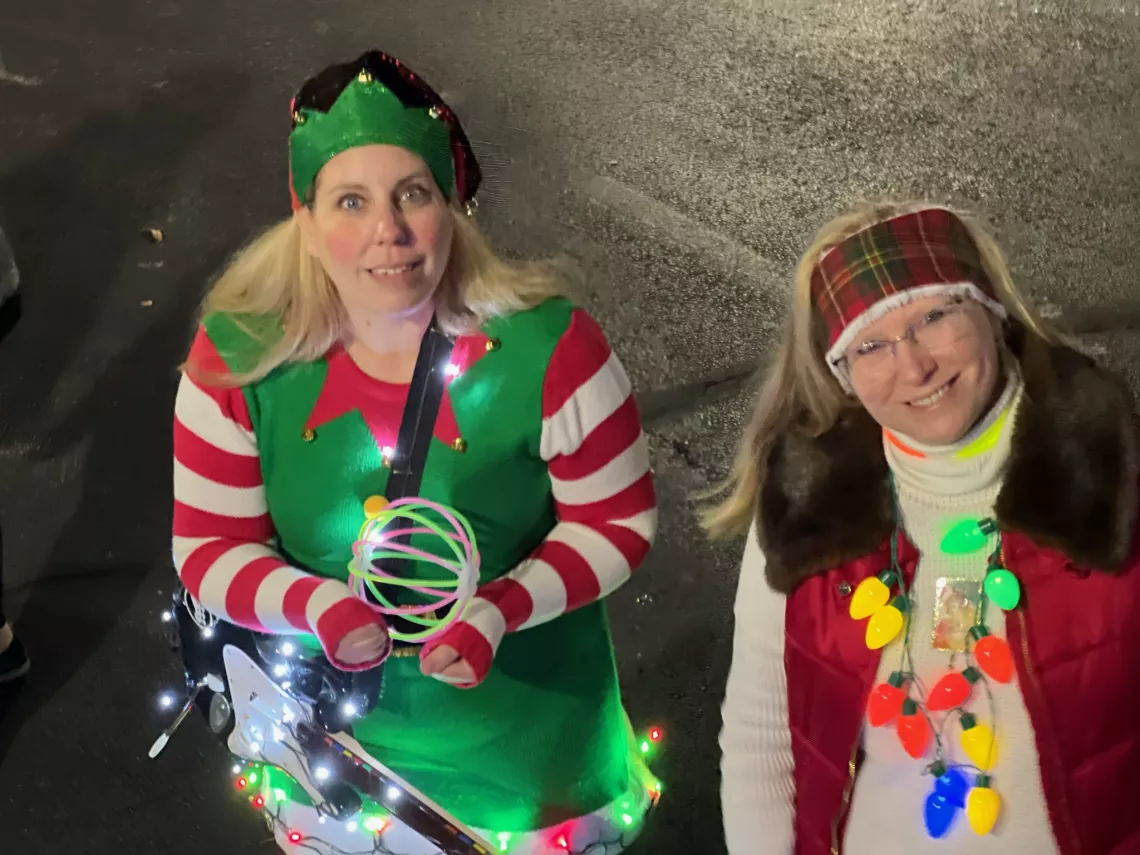 Two women with blonde hair wearing Christmas attire. One has a green elf hat and a green and red elf outfit. The other has string Christmas lights around her neck.