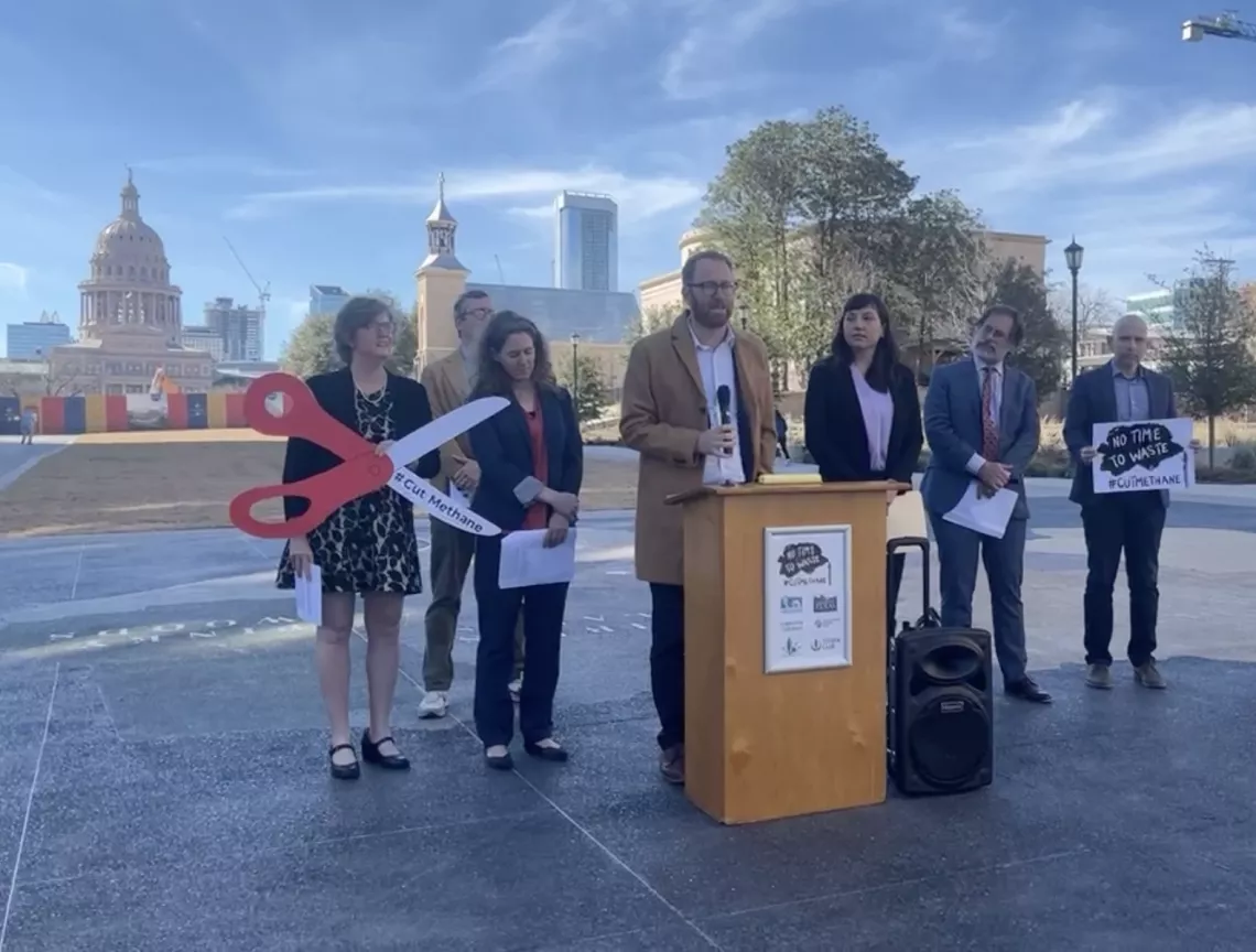 Seven people stand behind a podium with the Texas Capitol in the background. A woman holds a giant pair of scissors that say "Cut methane"