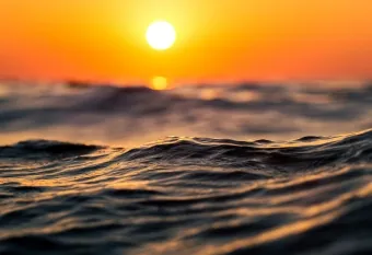 A bright yellow sun sets against a bright orange sky as ocean waves form in the foreground.