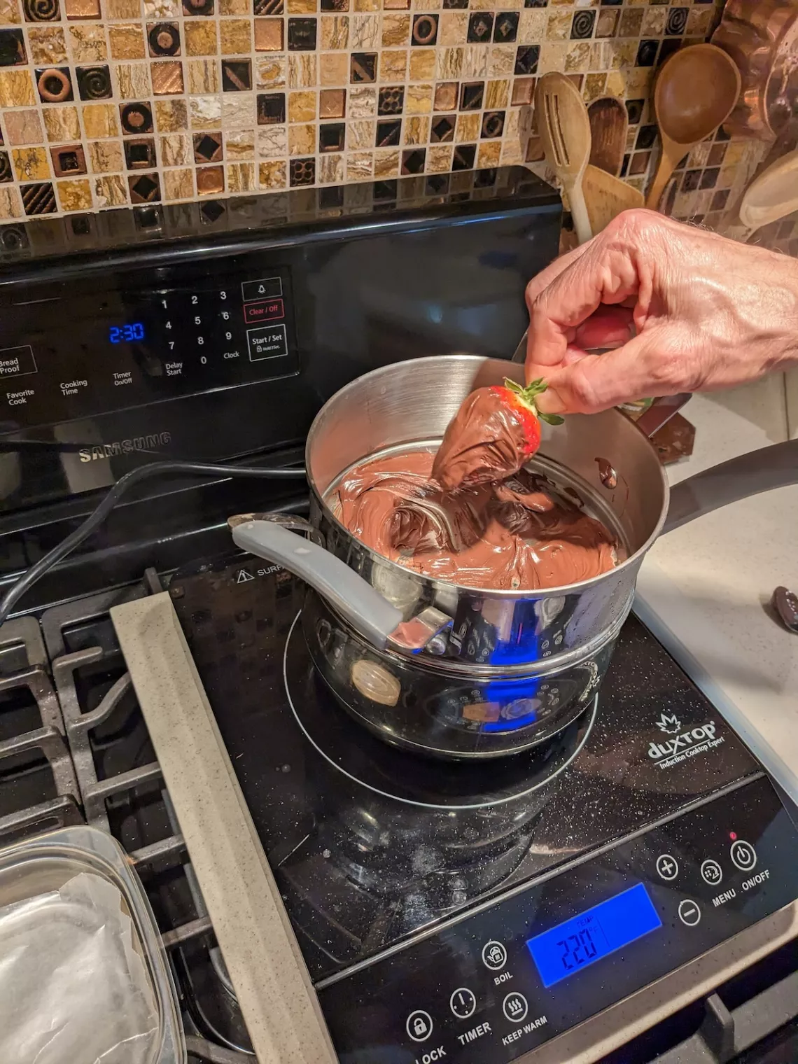 A hand dipping a strawberry into melted chocolate on a stove