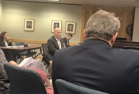 A bald man in a suit speaks into a microphone in a conference room. A woman with dark hair behind him appears to be concentrating hard. In the foreground is the back of a man in a suit jacket.