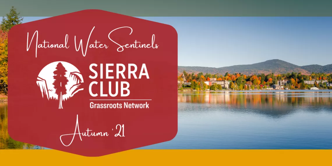A placid lake reflects blue sky and trees with autumn leaves.