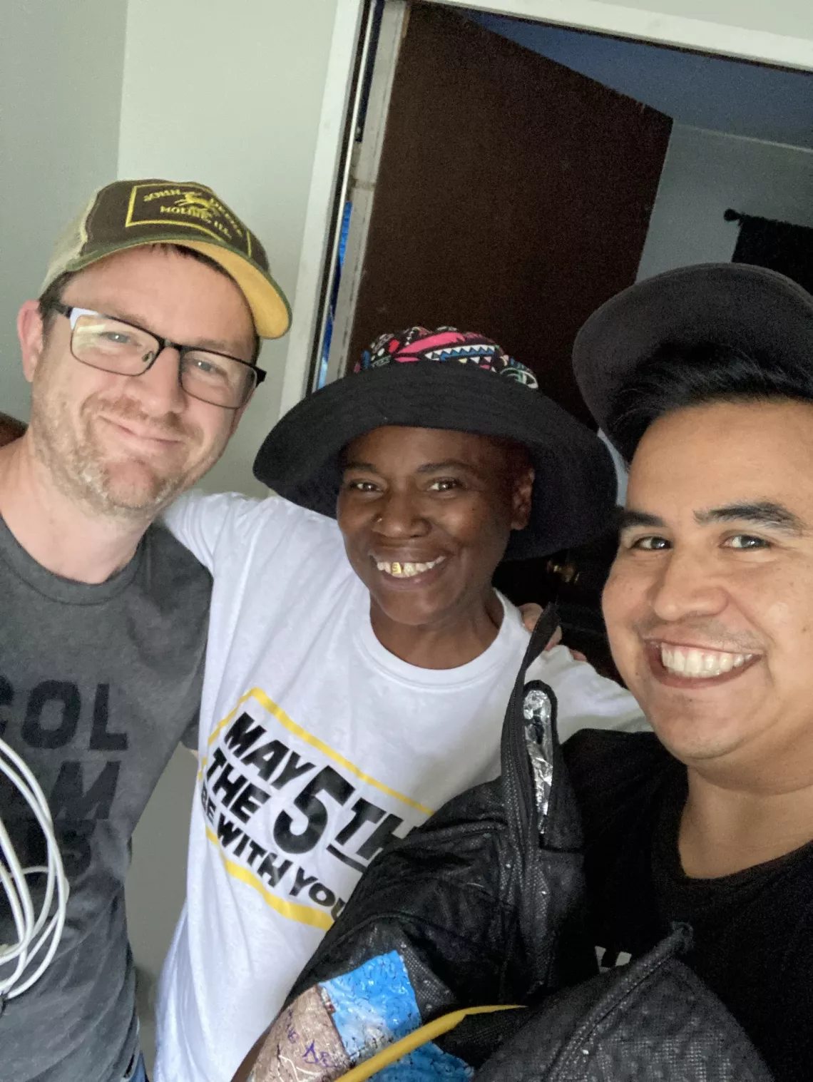 A tall white man, a Black woman in a hat, and a Latino man in a hat embrace.