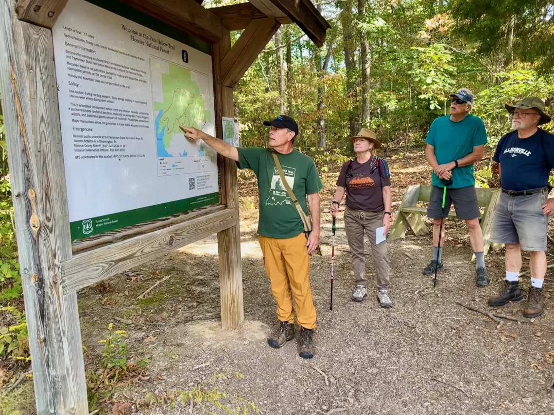 Four people at a trailhead looking at a map of the park. A couple of the folks have walking poles and they're all wearing hats to protect from the sun and bugs. The ground has some fallen brown leaves but the leaves on the trees behind them are green.