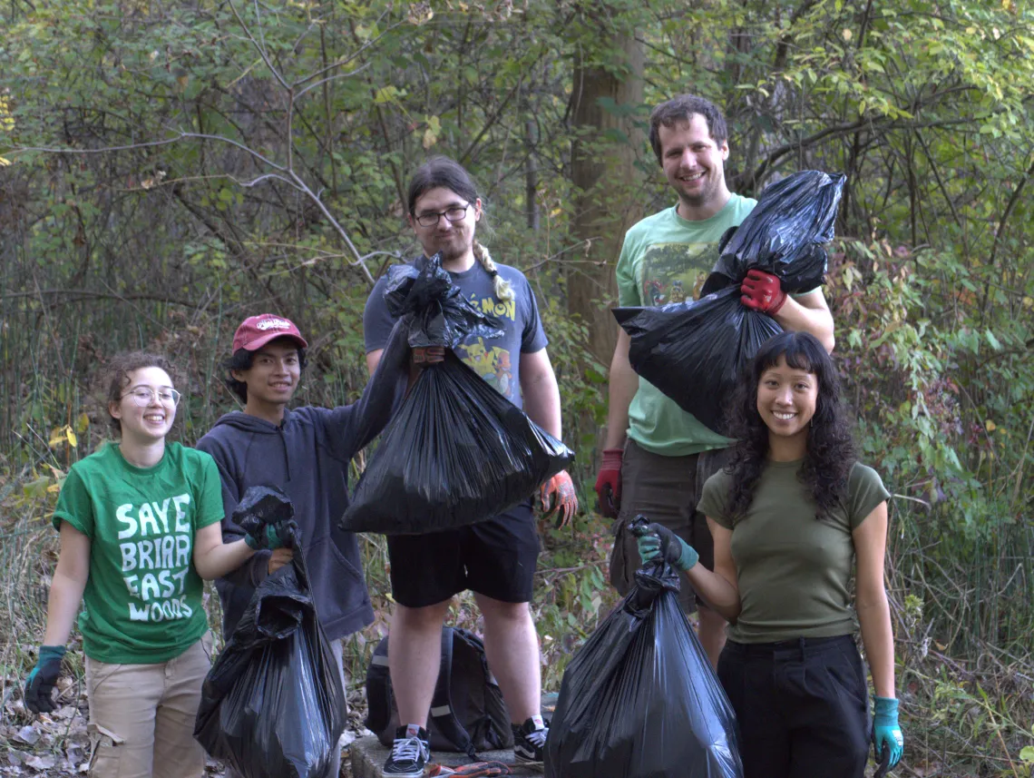 A group of five people outdoors with trees behind them. They're holding black trash bags. Some are wearing gloves, and one is wearing a green t-shirt with Save Briar East Woods in white letters.