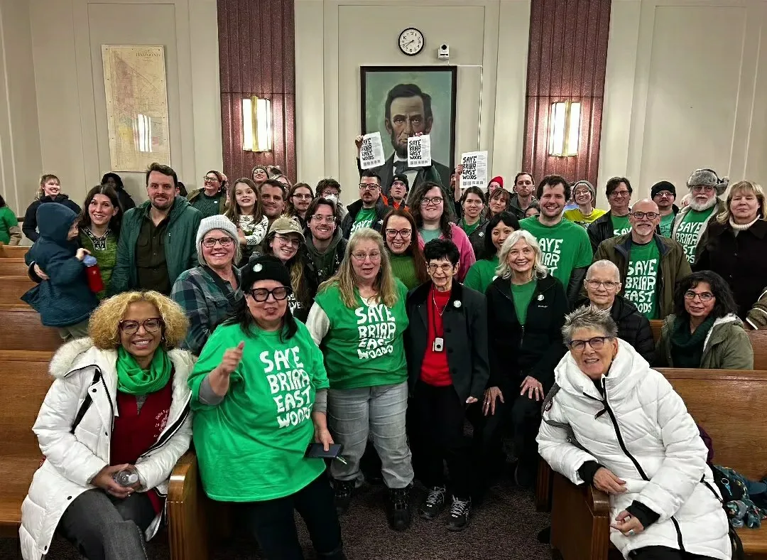 A large group of people indoors in an official building. Several of them are wearing green t-shirts that state Save Briar East Woods in white. Some are holding signs. They're all looking towards the camera for the photo.