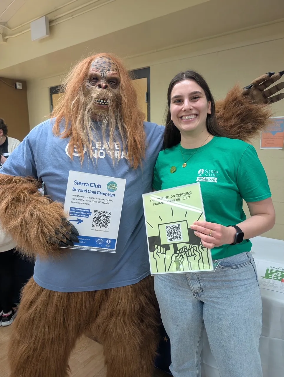 A person in a green t-shirt with the words Sierra Club organizer stood next to a person dressed as a very hairy mascot with a blue t-shirt. The mascot is holding a sign for a Sierra Club Beyond Coal campaign with a QR code, and the person in the green t-shirt is holding a sign saying to oppose Indiana senate bill 1007