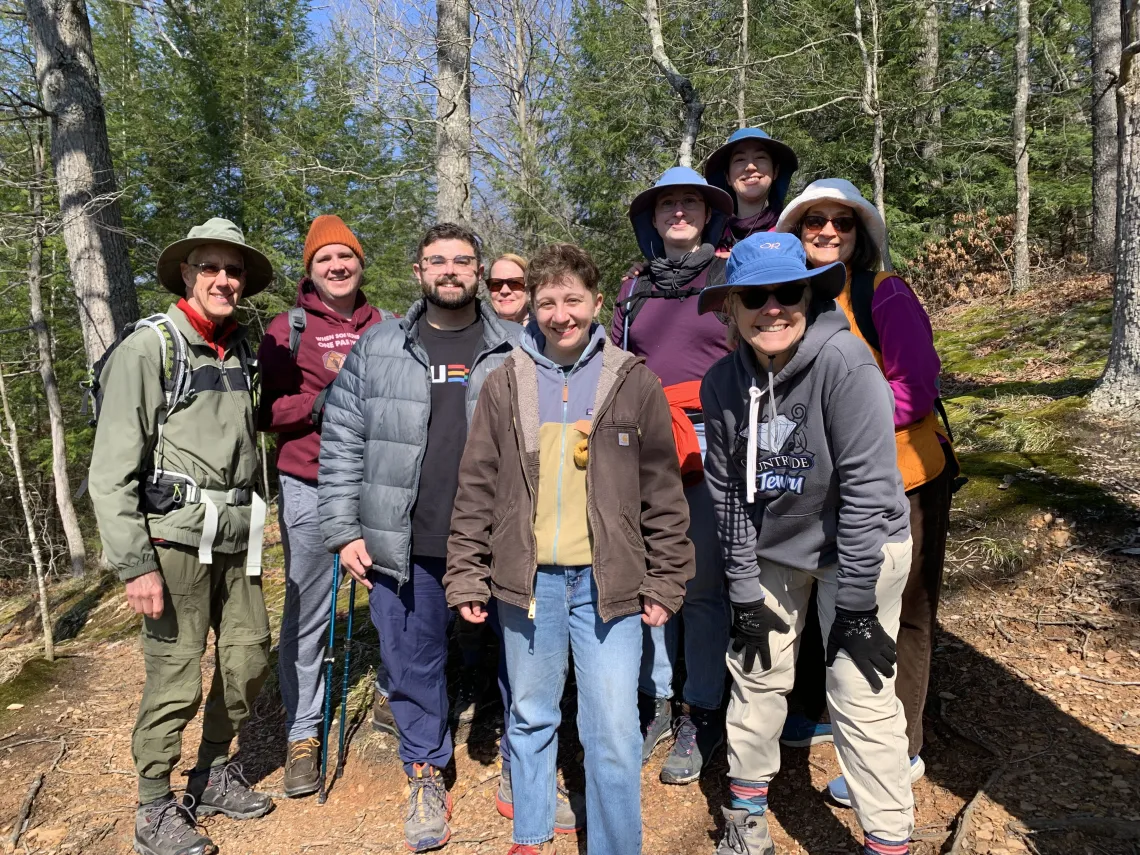 A group of nine people stood outdoors on a trail in a woods. It is a sunny day and there are pine trees with green needles behind them. Everyone in the group is smiling.