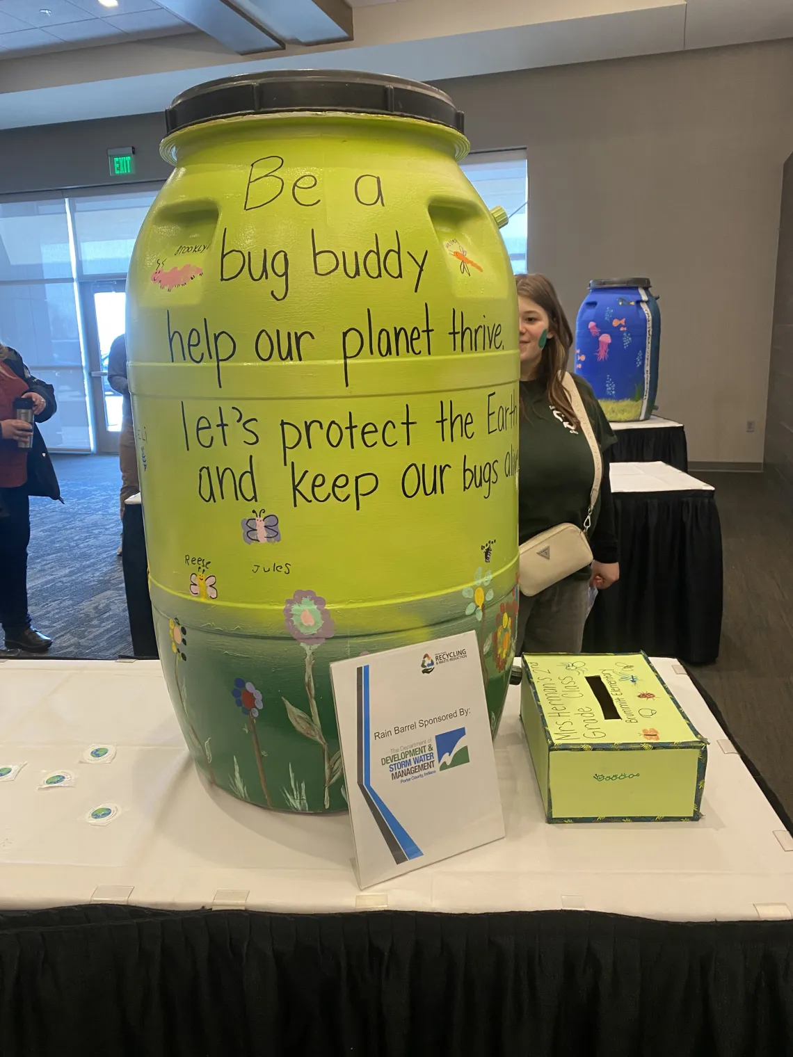 A large yellow rain barrel on a table with the words Be a bug buddy. Help our planet thrive. Let's protect the Earth and keep our bugs alive.