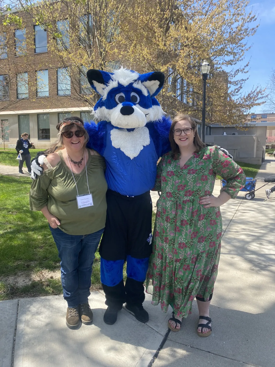 Two people stood either side of a blue and white furry animal mascot. They are both smiling. The person on the left is wearing a green top and blue jeans, and the person on the right is wearing a green dress with purple and white flowers. There is a building behind them and trees and blue sky.