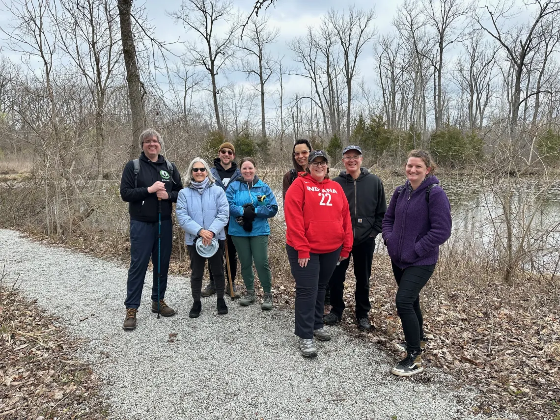 A group of 8 people stood outdoors on a gravel trail. There is a large pond behind them as well as bare trees, and brown leaves on the ground. They are dressed for a cool day.