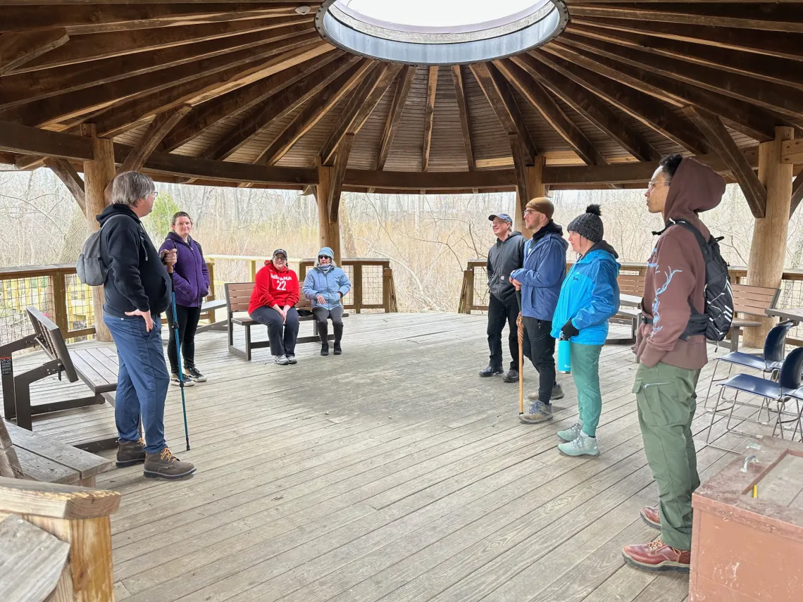A group of 8 people in an outdoor classroom, which is wood with a domed roof with a circular hole in the middle. Some are standing and some are seated on wooden benches.