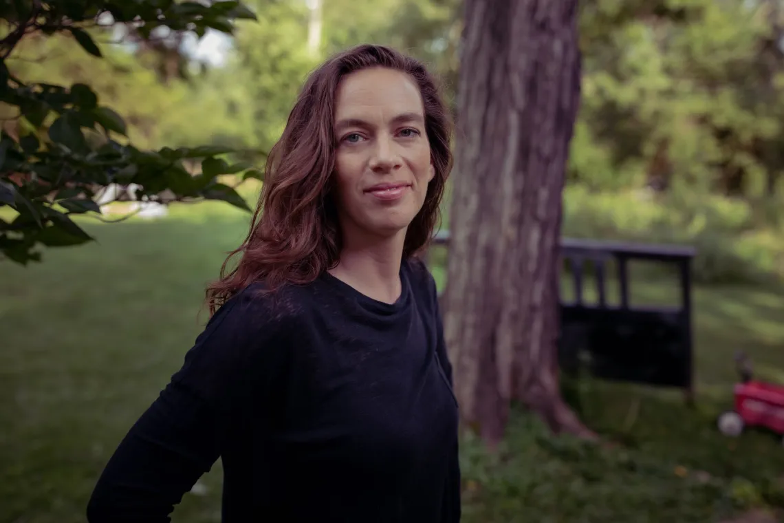 A woman with long brown hair stood outside in her yard - the background of trees and grass is slightly blurred in contrast with the person in the foreground. She is standing side on but looking towards the camera and is wearing a dark top with long sleeves.