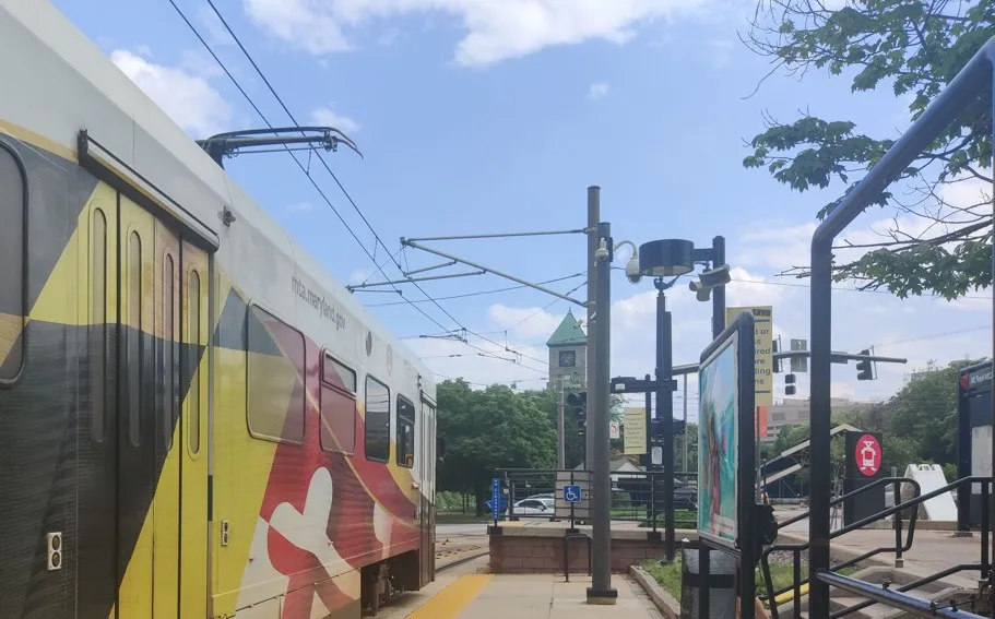 Light rail vehicle painted Maryland flag pulls up next to a station on sunny day with a clock tower building in the background