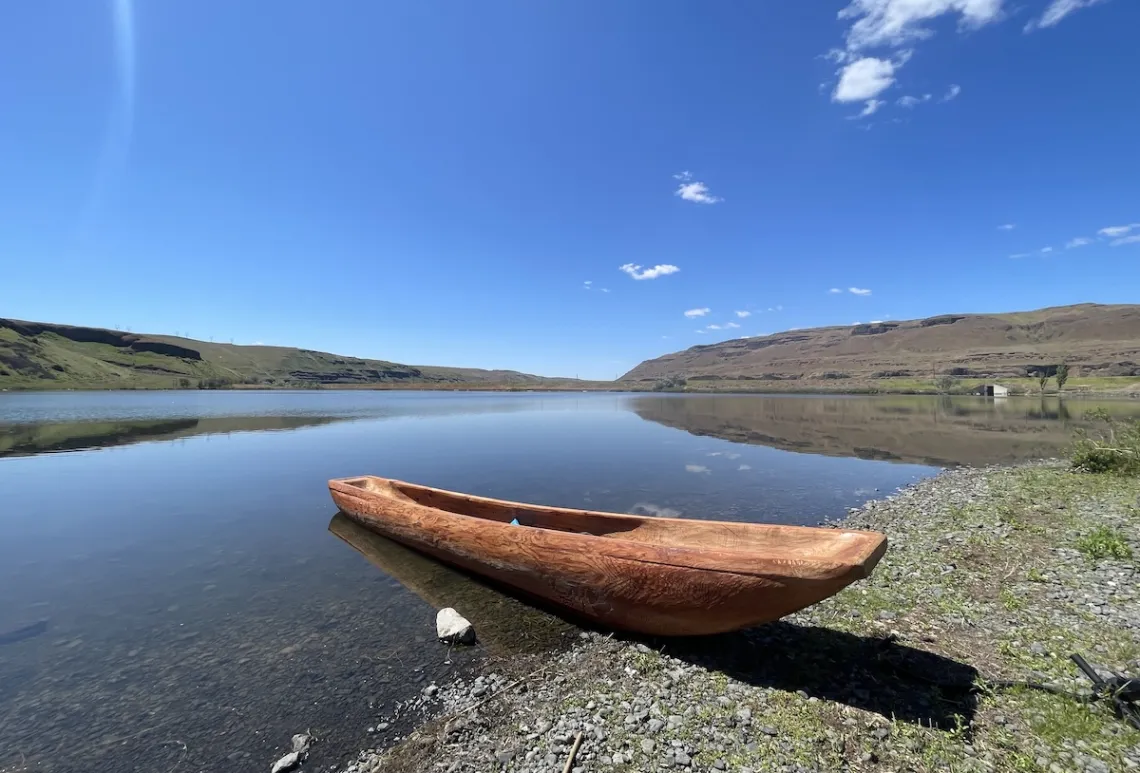 A carved wooden canoe sits alone and empty at the edge of a wide river