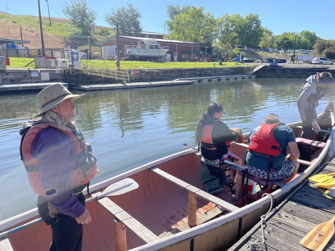 A man stands in the back of a canoe that's tied to a wooden dock. In the front of the canoe sit two passengers with their backs to the camera.