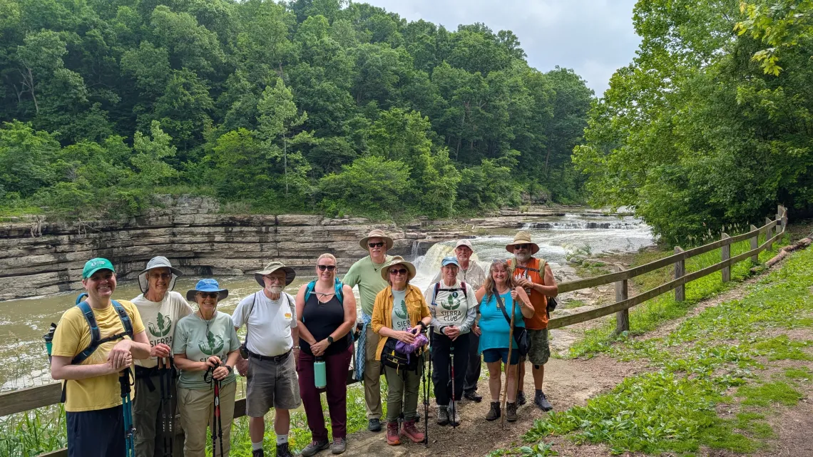A group of eleven people on a trail with a waterfall and green trees in the backround. Most are wearig sun hats and some have hiking polls. They're smiling.