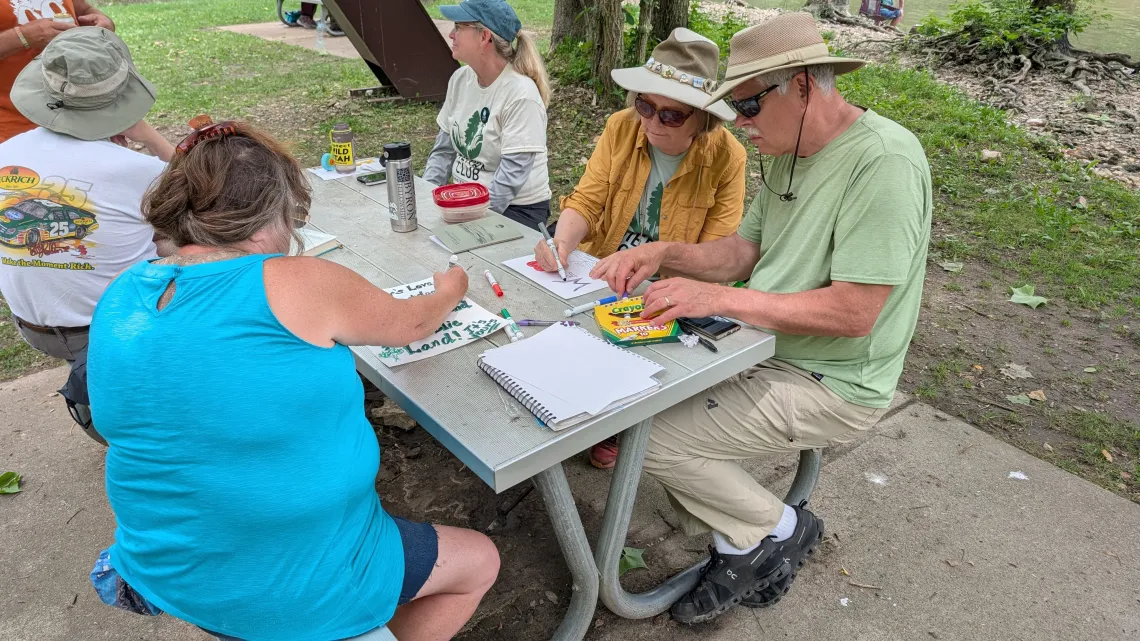 A group of five poeple sitting outdoors at a picnic table, making signs