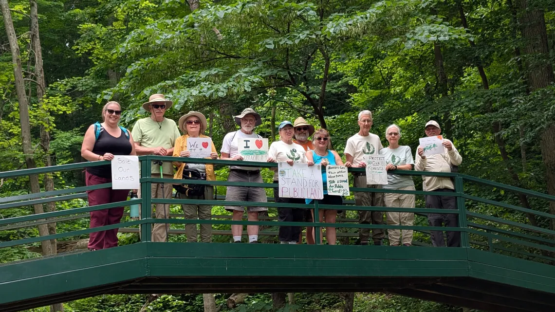 A group of ten people on a bridge with green trees in the background. Some are holding signs which say things like 'take a stand for public land' and 'save our forests'