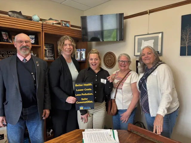 A group of five people stand in a line inside a small office holding a sign that says, "Better health, lower costs, less pollution, safer communities."