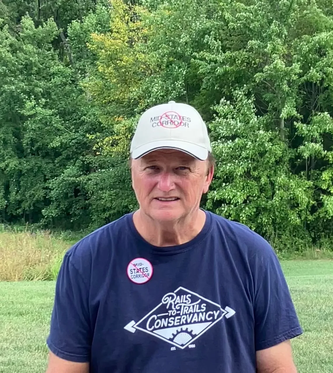 A man outdoors with green grass and green trees in the background. He has a white cap stating No Mid-States Corridor, a blue t-shirt, and a button also against the Mid-States Corridor