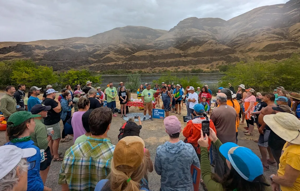 A large group of people stand next to a river while listening to a speaker, center in the photograph