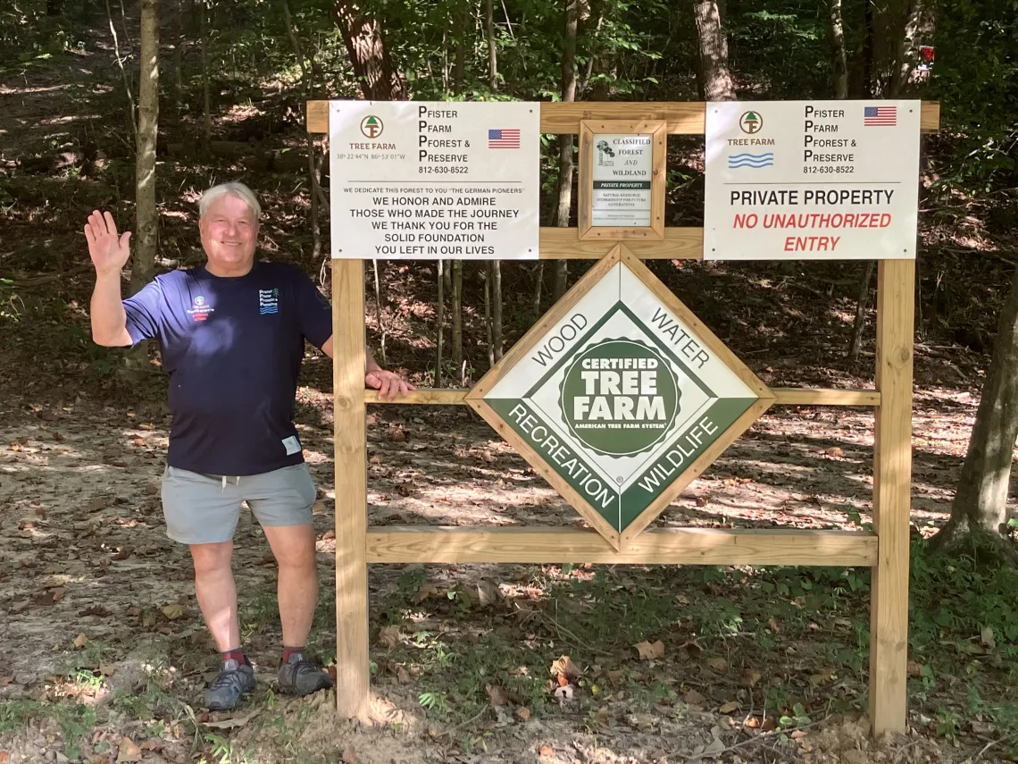 A person standing by a forest with a sign saying Certified Tree Farm - Wood Water Recreation Wildlife. He is wearing a blue tee, gray shorts, and is waving for the photo