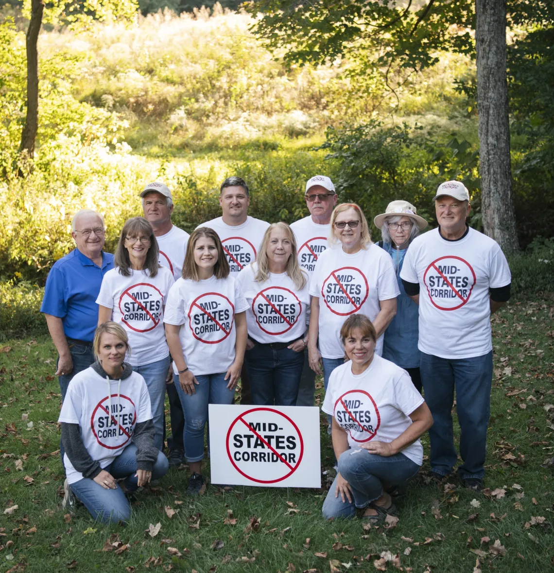A group of 12 people, most of them wearing white tees stating Stop the Mid-States Corridor. They are outdoors with green grass and some trees in the background. There is also a white Stop the Mid-States Corridor yard sign in the photo.