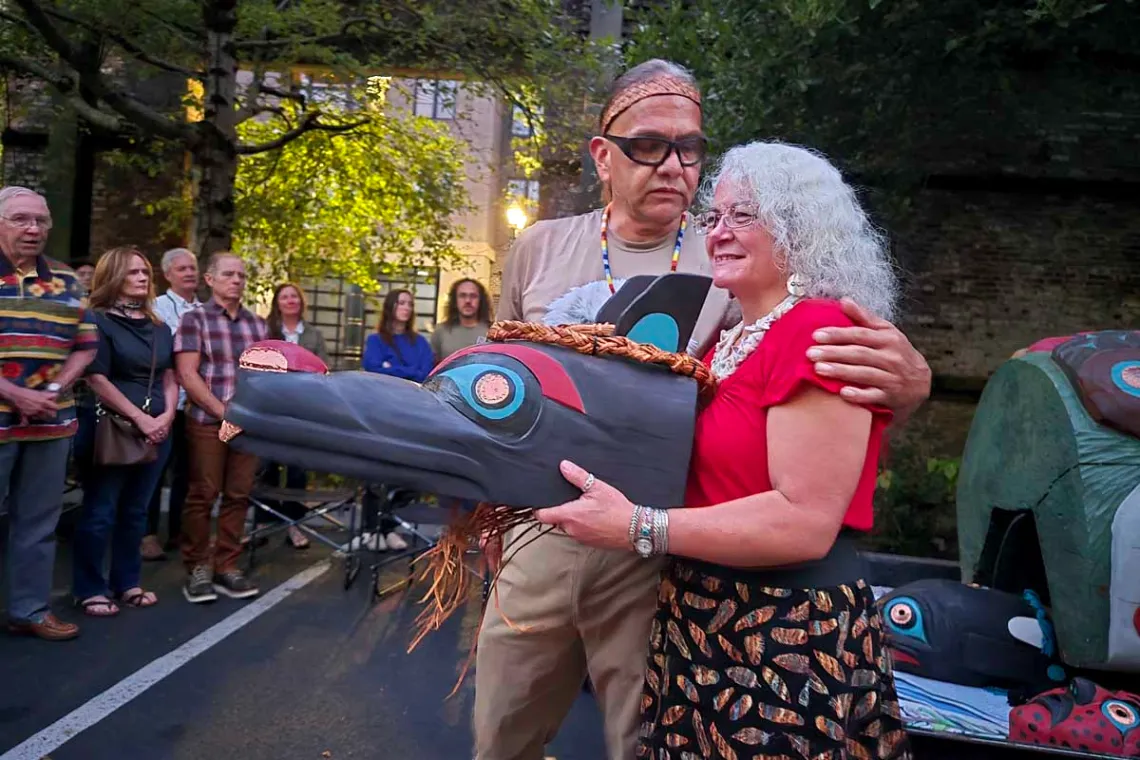 A man has his arm around a woman who holds a large cedar mask in front of her body
